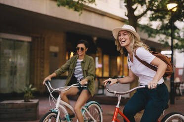 Smiling young woman cycling with her friend on city street. Two young women riding bicycles outdoors and having fun. - JLPSF25943