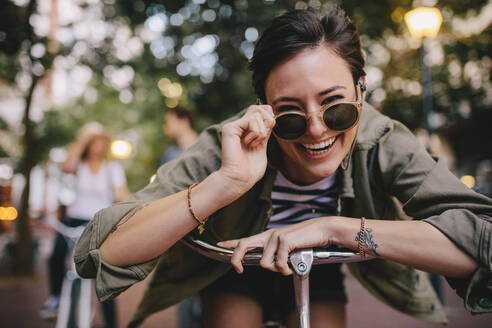 Fröhliche Frau mit Sonnenbrille, die sich im Freien an ihr Fahrrad lehnt. Frau genießt die Natur mit Freunden im Hintergrund. - JLPSF25946