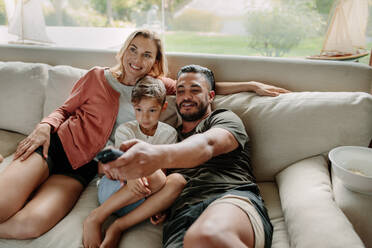 Family of three sitting on a sofa and watching TV together at home. Man with his son and wife relaxing on couch watching television. - JLPSF25954