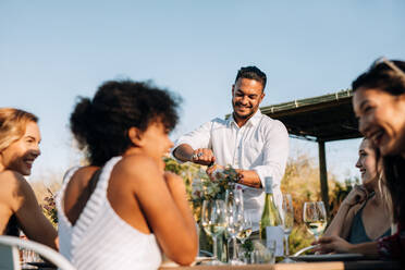 Mann öffnet Champagner mit Freunden, die während einer Party am Tisch sitzen. Multi-ethnische Millennials feiern eine Party im Freien in einem Restaurant. - JLPSF25975