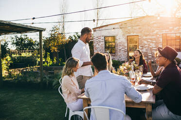 Ein Mann hält eine Rede vor einer Gruppe von Freunden auf einer Party. Junge Leute, die einen besonderen Anlass feiern, sitzen um einen Tisch in einem Gartenrestaurant. - JLPSF25976