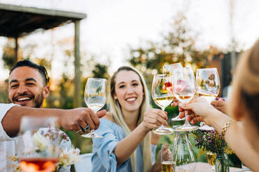 Eine Gruppe von Freunden stößt mit Wein an und amüsiert sich im Freien in einem Restaurant, wobei die Weingläser im Mittelpunkt stehen. - JLPSF25986
