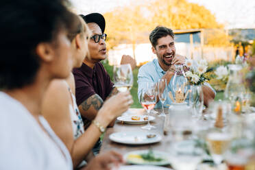 Junger Mann lacht, während er mit Freunden auf einer Party im Freien sitzt. Freunde genießen eine Sommermahlzeit in einem Restaurant im Freien. - JLPSF25987