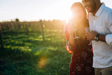 Lächelndes Paar, das mit einem Glas Wein an einem Sommertag spazieren geht. Mann und Frau mit einem Getränk lachend im Freien auf einem Weinberg. - JLPSF25989