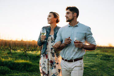 Mann und Frau mit einem Getränk im Freien spazieren gehend. Paar zusammen mit einem Glas Wein in einem Weinberg. - JLPSF25991