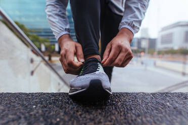 Männlicher Läufer beim Binden der Schnürsenkel auf der Treppe eines Gebäudes. Nahaufnahme eines Sportlers mit Laufschuh. - JLPSF26015