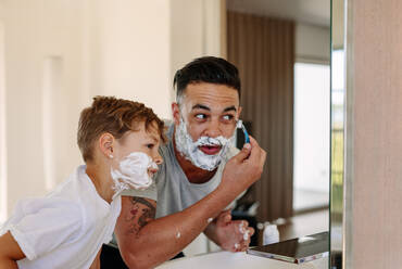 Father and son shaving together at home bathroom. Young man and little boy with shaving foam on their faces are shaving and looking into the mirror. - JLPSF26047