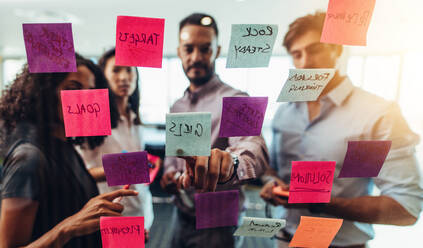 Business investors discussing work looking at the information on sticky notes stuck in office. Colleagues standing in front of post-it notes stuck on glass and discussing. - JLPSF26074