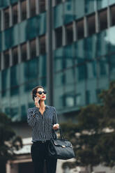 Asian businesswoman walking down the city street and talking on mobile phone. Female business professional walking outside with an office building in background. - JLPSF26077