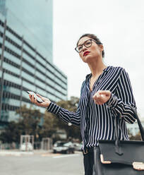 Female business executive holding cell phone, hailing taxi. Asian businesswoman waiting for taxi on the city street. - JLPSF26081