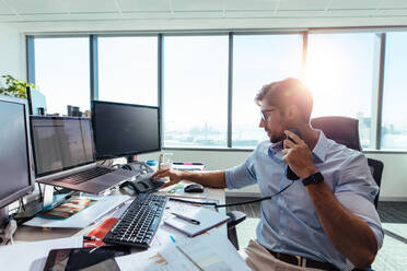 Geschäftsmann, der in seinem Büro mit Computern und Geschäftspapieren auf dem Tisch arbeitet. Junger Unternehmer, der einen Anruf mit einem Telefon an seinem Bürotisch tätigt. - JLPSF26083