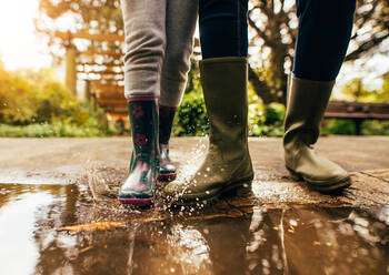 Low section view of mother and daughter feet wearing gum boots standing water puddle on road. Close up shot of mother playing with daughter in the pool of water on street. - JLPSF26087