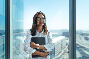 Glückliche Geschäftsinvestorin in ihrem Büro in einem Hochhaus mit Blick auf die Stadtlandschaft. Geschäftsfrau mit Brille hält eine Dokumentenmappe in der Nähe des Fensters im Büro. - JLPSF26093