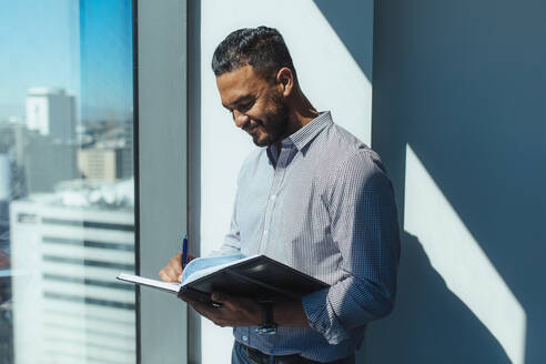 Junger Geschäftsmann schreibt Notizen in Tagebuch am Fenster im Büro stehend. Lächelnder Mann bei der Büroarbeit stehend im Sonnenschein am Fenster eines Hochhaus-Bürogebäudes mit Blick auf die Stadtlandschaft. - JLPSF26097