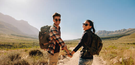 Schönes junges Paar beim Wandern im Naturschutzgebiet. Junger Mann und Frau halten sich an den Händen und schauen zurück, während sie an einem Sommertag wandern. - JLPSF26107