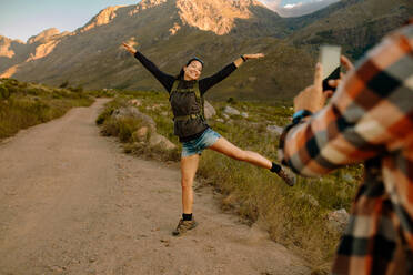 Aufgeregte junge Frau, die während einer Wanderung in der Natur für ein Foto posiert. Mann, der seine Freundin beim Wandern in der Natur im Sommerurlaub fotografiert. - JLPSF26119