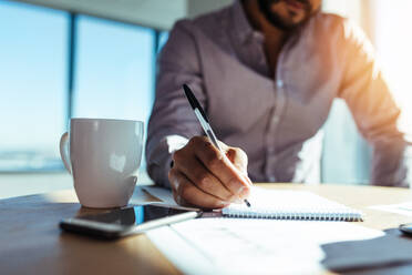 Geschäftsinvestor schreibt in einen Notizblock mit einer Kaffeetasse auf dem Schreibtisch. Nahaufnahme der Hand eines Geschäftsmannes, der einen Stift hält und sich Notizen macht. - JLPSF26129