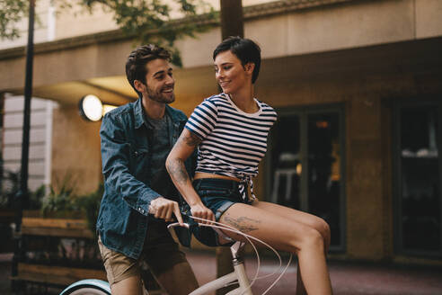 Glücklicher junger Mann fährt Fahrrad mit einer Frau, die auf dem Lenker sitzt. Romantisches Paar, das eine Fahrradtour in der Stadt genießt. - JLPSF26136