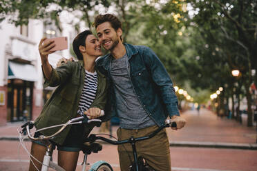 Romantisches Paar mit Fahrrädern, das ein Selfie macht. Frauen, die ein Selfie machen und ihren Freund auf die Wange küssen. - JLPSF26140