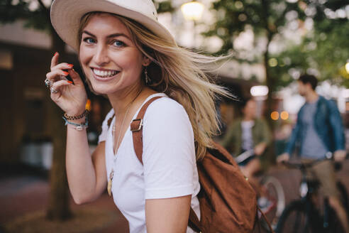 Nahaufnahme einer attraktiven jungen Frau mit Freunden auf ihren Fahrrädern im Hintergrund. Lächelnde Frau mit Hut und Tasche, die im Freien steht. - JLPSF26152