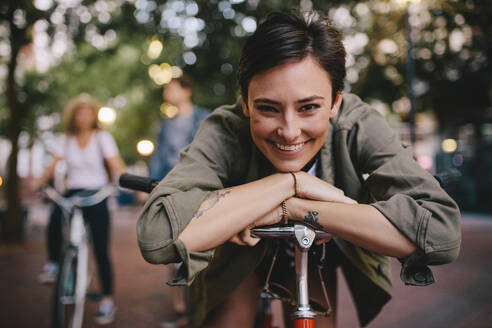 Porträt einer glücklichen Frau, die sich auf ihr Fahrrad stützt, mit Freunden im Hintergrund. Eine Frau genießt einen Tag in der Stadt mit ihrem Fahrrad und ihren Freunden. - JLPSF26153