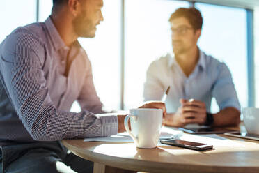 Nahaufnahme einer Kaffeetasse mit unscharfem Bild von zwei Geschäftsleuten, die am Tisch sitzen. - JLPSF26167