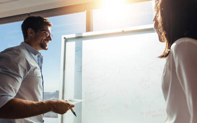 Geschäftsmann schreibt Ideen auf Whiteboard, während er die Arbeit mit einer Kollegin bespricht. Geschäftsinvestoren diskutieren im Sitzungssaal mit Sonnenschein im Hintergrund. - JLPSF26176