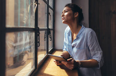 Woman standing by window with a digital tablet. Female business professional looking outside the window with tablet computer in hand. - JLPSF26190