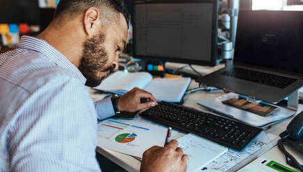 Geschäftsmann sitzt an seinem Tisch und analysiert Daten und Diagramme. Mann arbeitet an seinem Schreibtisch mit Computern, Notizen und Präsentationsunterlagen. - JLPSF26191
