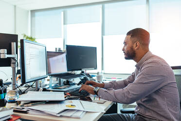 Ein Mann arbeitet an seinem Schreibtisch mit Computern, Notizen und Präsentationsunterlagen. Ein Geschäftsmann analysiert Diagramme und Tabellen für die Entscheidungsfindung. - JLPSF26192