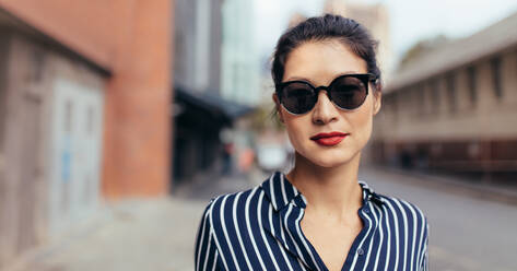 Portrait of young woman wearing sunglasses walking outside on the city street. Asian female model walking outdoors and looking at camera. - JLPSF26193