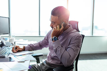 Unternehmer macht sich Notizen, während er am Handy spricht. Mann sitzt an seinem Schreibtisch und bespricht seine Arbeit am Telefon. - JLPSF26195