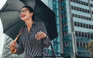 Young smiling business woman walking down the street with umbrella. Happy female business professional with umbrella outdoors. - JLPSF26209