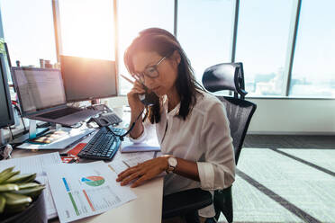 Frau Unternehmer Blick auf Geschäftspapiere während des Gesprächs über das Telefon. Frau sitzt an ihrem Schreibtisch im Büro arbeiten mit Papieren, während vor Desktop-Computern sitzen. - JLPSF26215