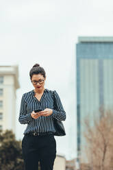 Junge asiatische Geschäftsfrau, die im Freien spazieren geht und ihr Handy benutzt. Weibliche Geschäftsfrau, die im Freien spazieren geht und mit ihrem Handy SMS schreibt. - JLPSF26217
