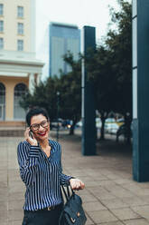Lächelnde junge asiatische Geschäftsfrau, die im Freien spazieren geht und mit einem Mobiltelefon spricht. - JLPSF26222