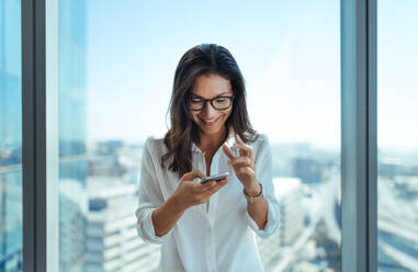 Junge Frau, die lächelt und gestikuliert, während sie ihr Mobiltelefon benutzt. Junge Frau in ihrem Büro in einem Hochhaus mit Blick auf die Stadtlandschaft. - JLPSF26228