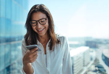 Frau mit Brille lächelt, während sie auf ihr Mobiltelefon schaut. Junge Geschäftsfrau, die ein Mobiltelefon für geschäftliche Kommunikation benutzt. - JLPSF26230