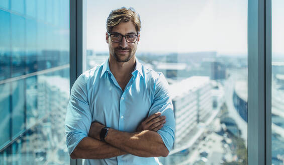 Nahaufnahme eines lächelnden Geschäftsmannes, der am Fenster seines Büros steht. Ein junger Mann mit Brille und verschränkten Armen steht in einem Büro. - JLPSF26235