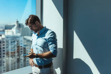 Junger Geschäftsmann, der am Fenster stehend auf sein Mobiltelefon schaut. Ein Mann in Formation steht am Fenster eines Bürohochhauses mit Blick auf die Stadtlandschaft. - JLPSF26242
