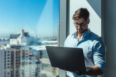 Junger Geschäftsmann, der in einem Büro an einem Laptop arbeitet. Mann mit Laptop am Fenster eines Bürohochhauses mit Blick auf die Stadtlandschaft. - JLPSF26243