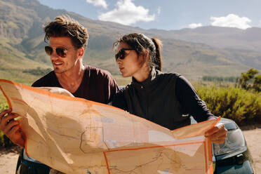 Young couple standing next to car and looking at map to find best route. Young man and woman holding a route map on a road trip. - JLPSF26271