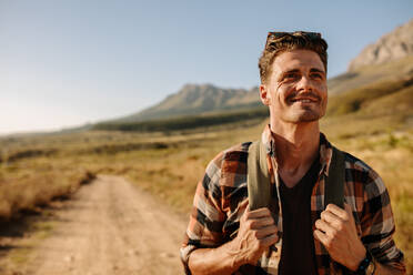 Lächelnder junger Mann mit Rucksack auf Landpartie, der wegschaut. Außenaufnahme eines männlichen Wanderers auf einem Wanderweg. - JLPSF26290