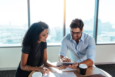 Smiling businessman and businesswoman in a discussion sitting at a table in office. Business colleagues using a tablet pc while discussing business financials. - JLPSF26304