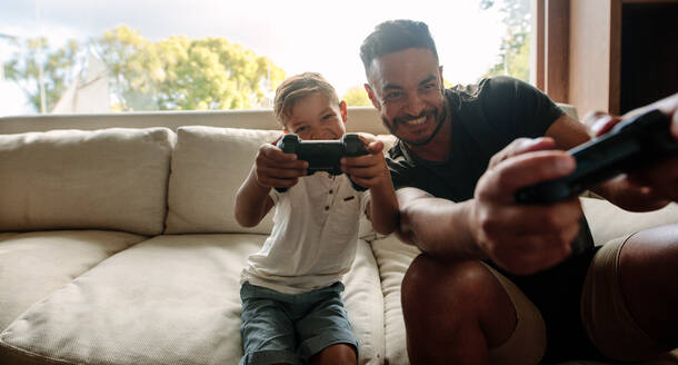 Porträt von Vater und Sohn, die im Wohnzimmer ein Videospiel spielen. Ein lächelnder junger Mann und ein kleiner Junge spielen zu Hause ein Videospiel. - JLPSF26305