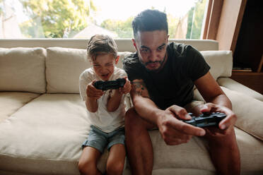 Father and son with video game controllers sitting on sofa. Young family having fun playing video games at home. - JLPSF26306