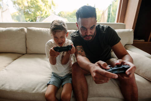 Vater und Sohn mit Videospiel-Controllern auf dem Sofa sitzend. Junge Familie hat Spaß beim Videospielen zu Hause. - JLPSF26306
