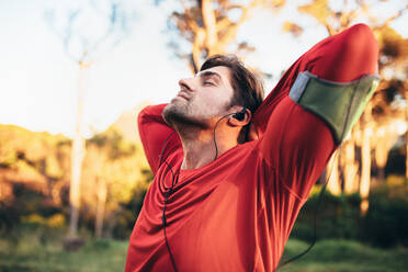 Ein Läufer macht Atemübungen, während er in einem Park Musik hört. Ein Mann entspannt sich mit geschlossenen Augen nach dem Training. - JLPSF26314
