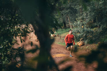 Athlete running on mud track in a park. Man running in park seen through dense trees. - JLPSF26316