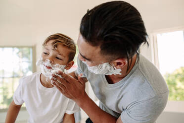 Mann trägt Rasierschaum auf das Gesicht seines Sohnes im Badezimmer auf. Vater und Sohn rasieren sich gemeinsam im Badezimmer. - JLPSF26346